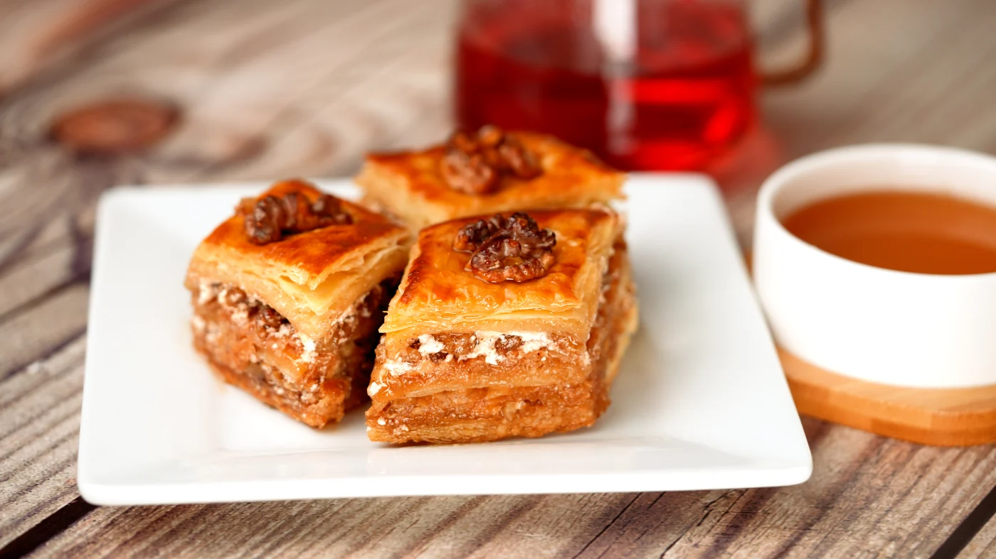 Baklava slice. Three golden square layered pastries with glossy tops and small brown decorative pieces, arranged on a white plate.