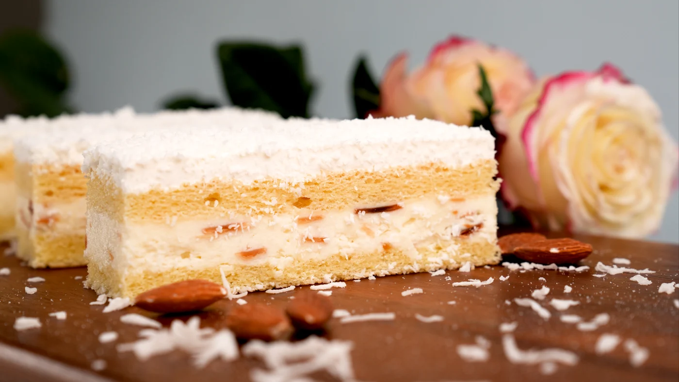 Coconut slice. Rectangular pale cake slice with white flakes on top and creamy-looking layers containing small pink pieces.