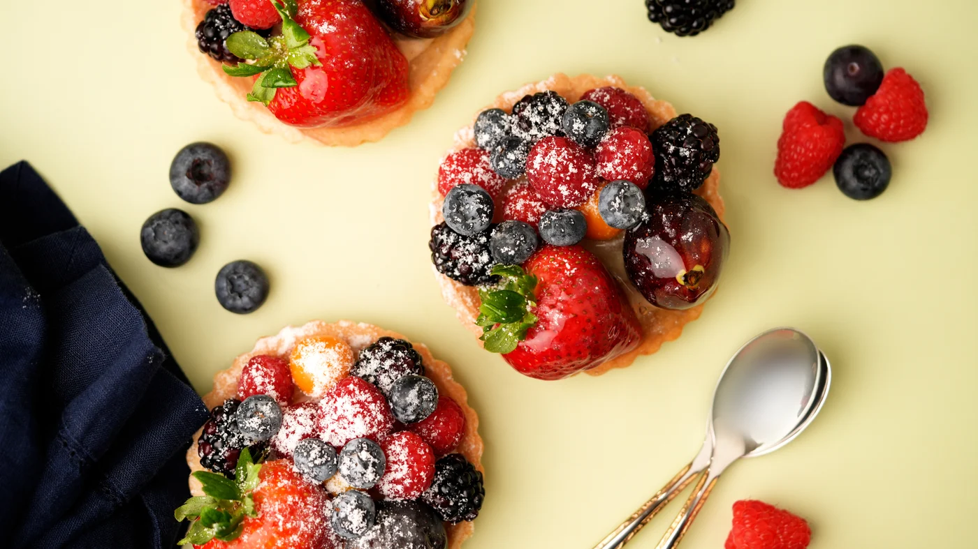 Fruit tart. Small round golden tarts topped with strawberries, blueberries, raspberries, and blackberries, viewed from above with a light white dusting.
