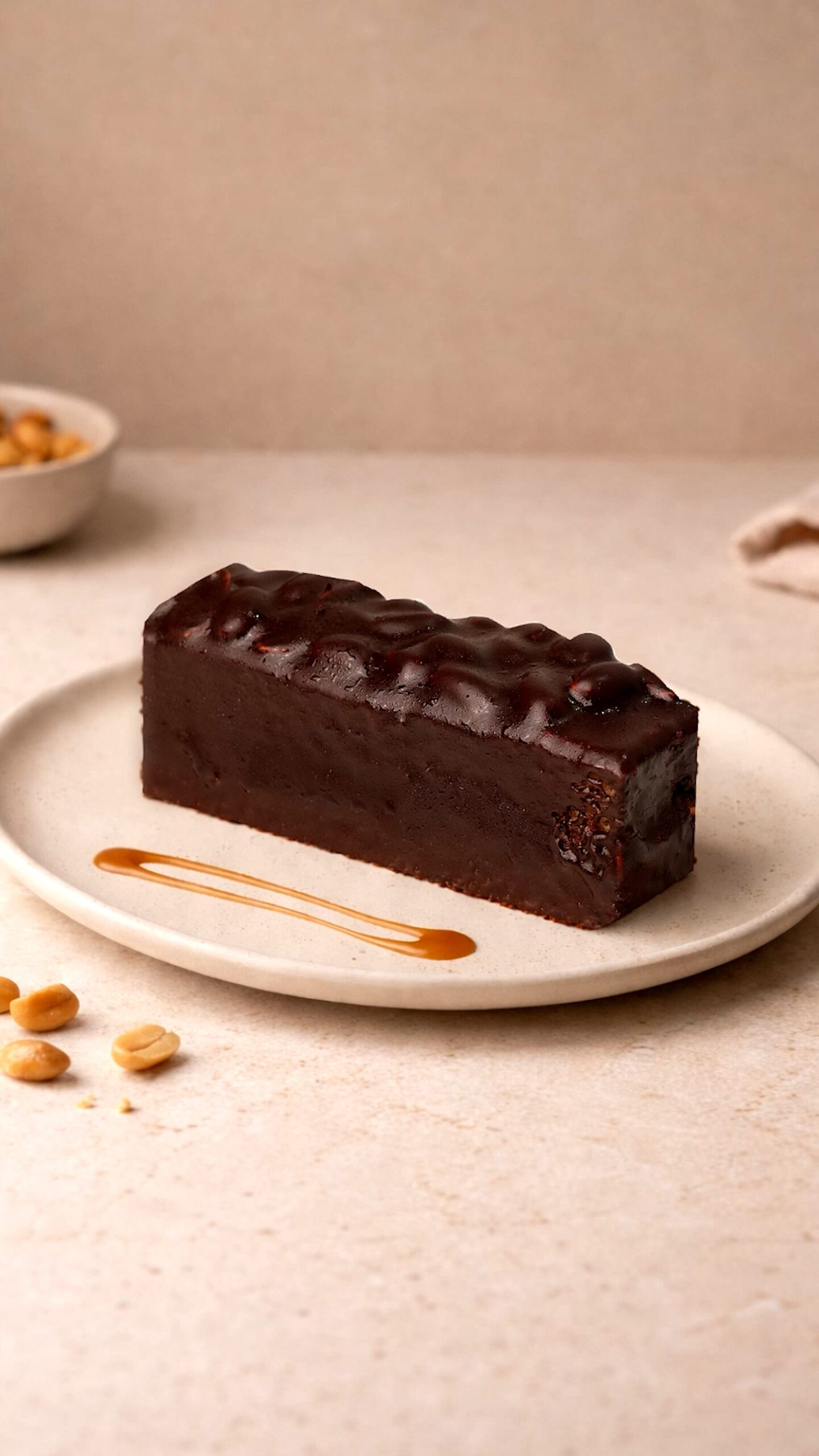 Snickers slice. Rectangular dark brown cake slice with glossy curled piping across the top, served on a pale plate.